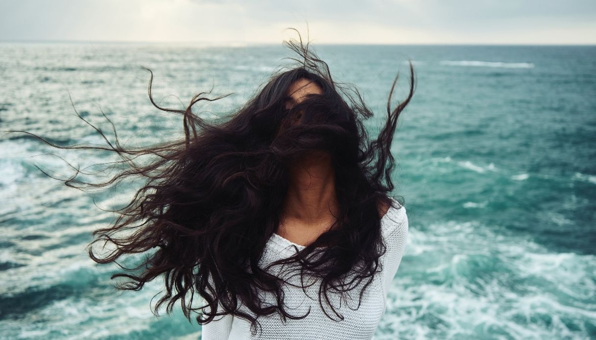 ragazza mora con capelli lunghi mossi dal vento davanti alla faccia con sfondo di mare mosso