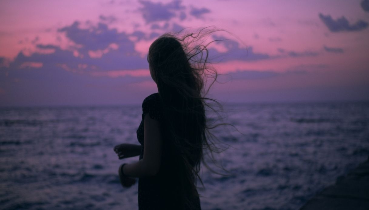 ombra di ragazza dai capelli lunghi al vento al tramonto sul mare con nuvole e cielo viola e rosa