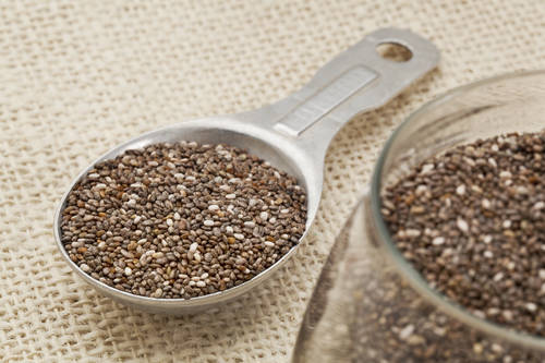chia seeds in glass jar and on measuring aluminum tablespoon against burlap background, focus on the spoon