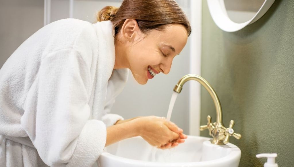Donna sorridente si lava la faccia in bagno con acqua fresca
