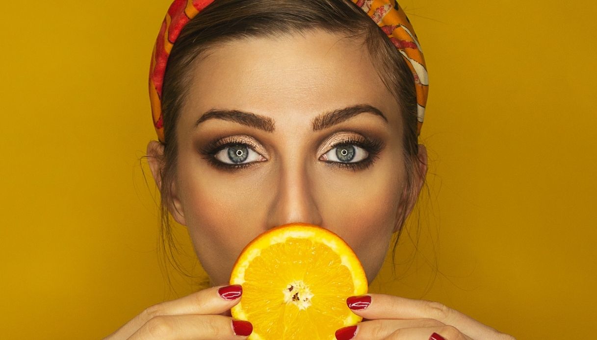 donna con occhi azzurri e trucco evidente si copre la bocca con una fetta di arancia