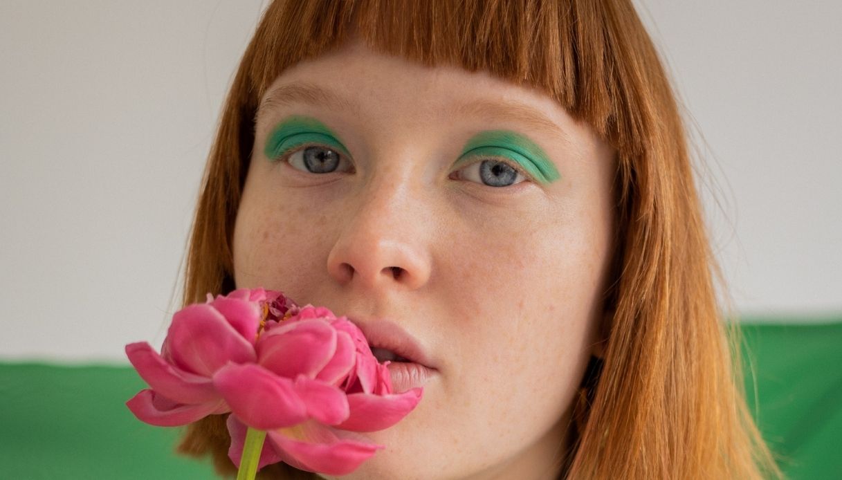 ragazza capelli rossi con frangia con fiore rosa davanti alla bocca e ombretto verde