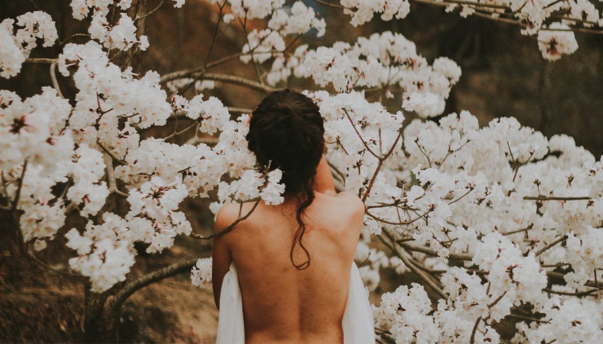 donna mora con capelli raccolti schiena. nuda con asciugamano bianco e sfondo di fiori bianchi