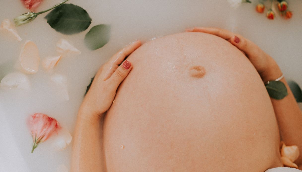 primo piano pancia donna incinta con mani sulla pancia e smalto rosso in vasca di acqua lattiginosa