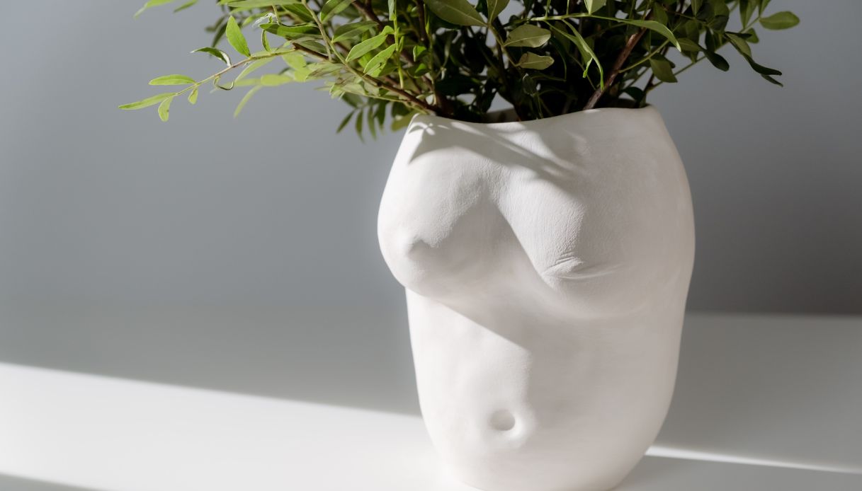 vaso di fiori bianco con petto di donna con seno e ombelico al cui interno ci sono piante verdi è appoggiato su un tavolo bianco