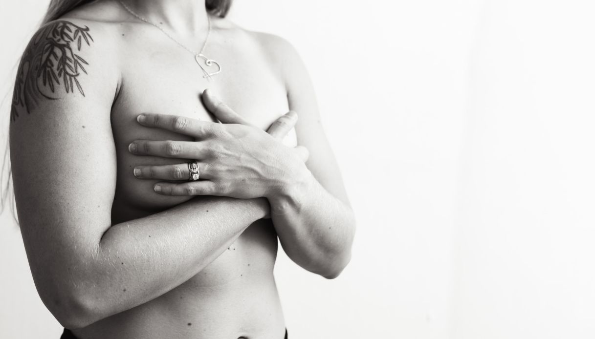 donna nuda con tatuaggio sulla spalla copre il seno con le mani incrociate davanti