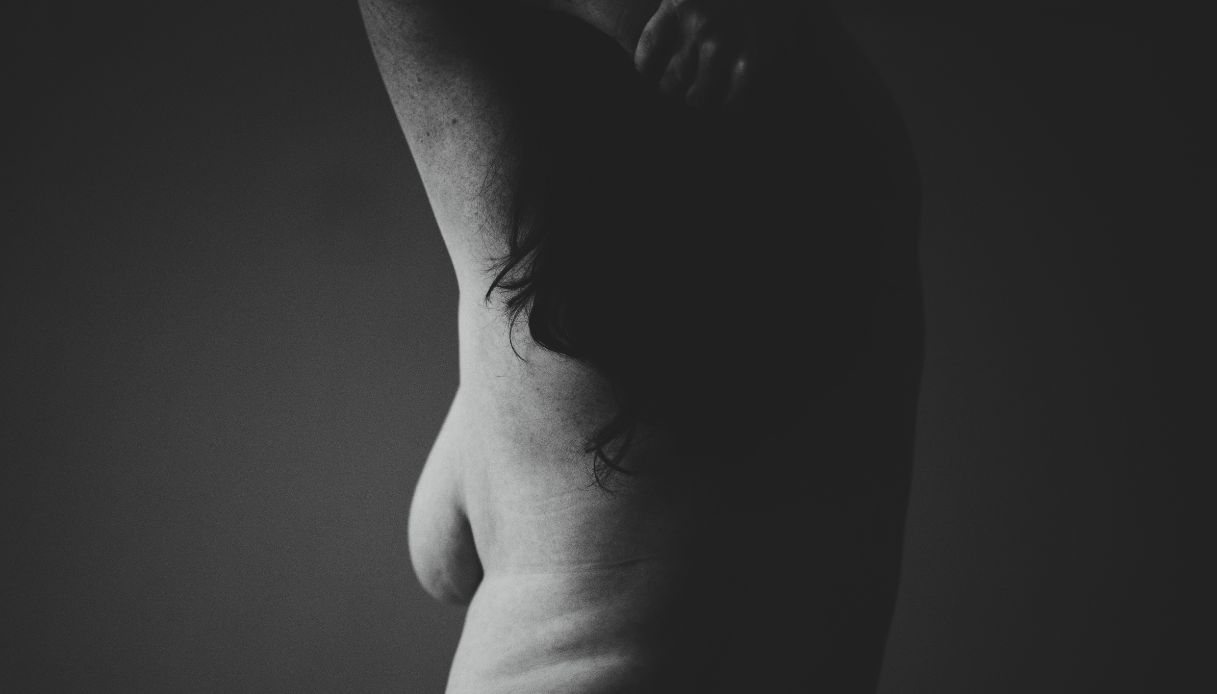 donna nuda morbida di spalle con braccia tirate su, capelli lunghi e laterale del seno che si intravede