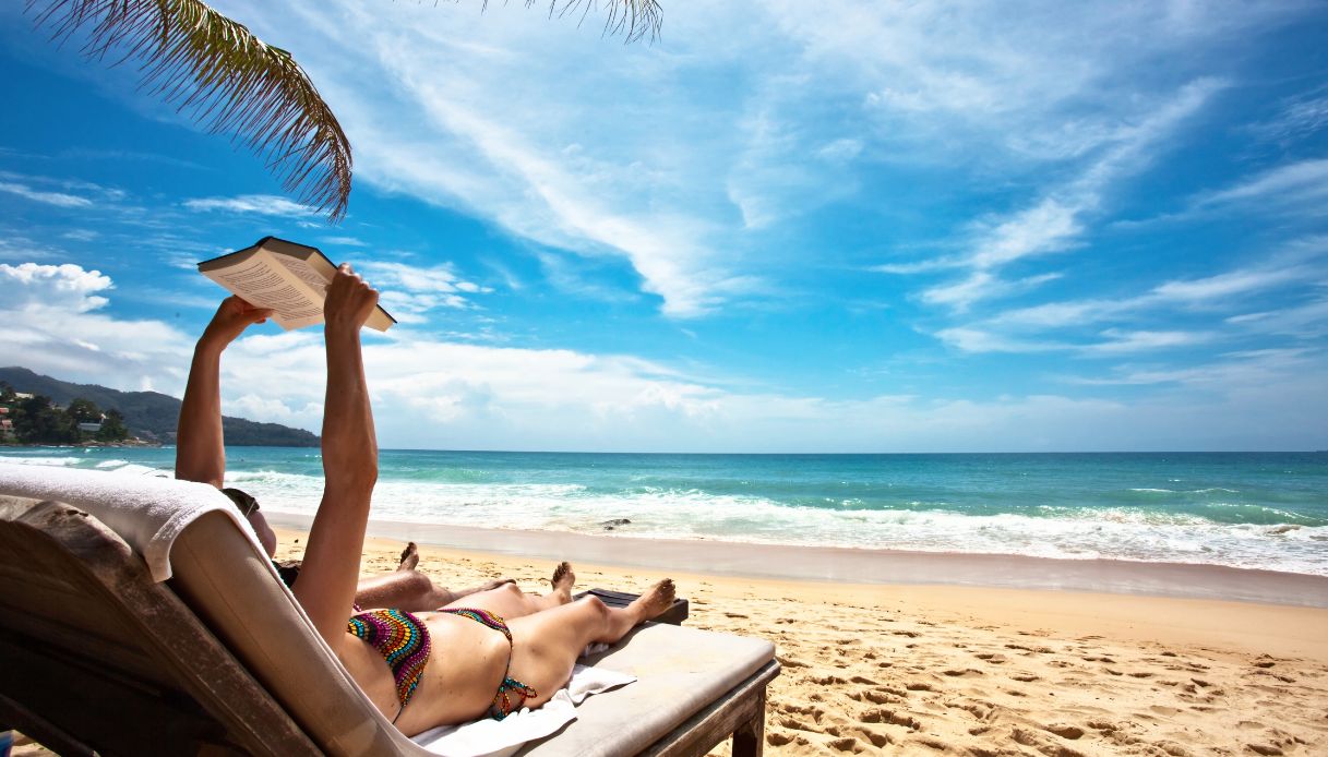 donna sdraiata su lettino prende il sole leggendo un libro con sfondo spiaggia e mare