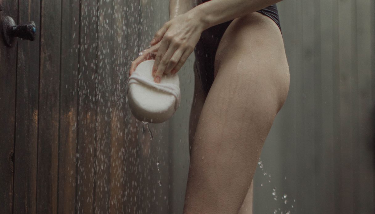 primo piano gambe donna con costume nero che fa doccia con spugna bianca in mano