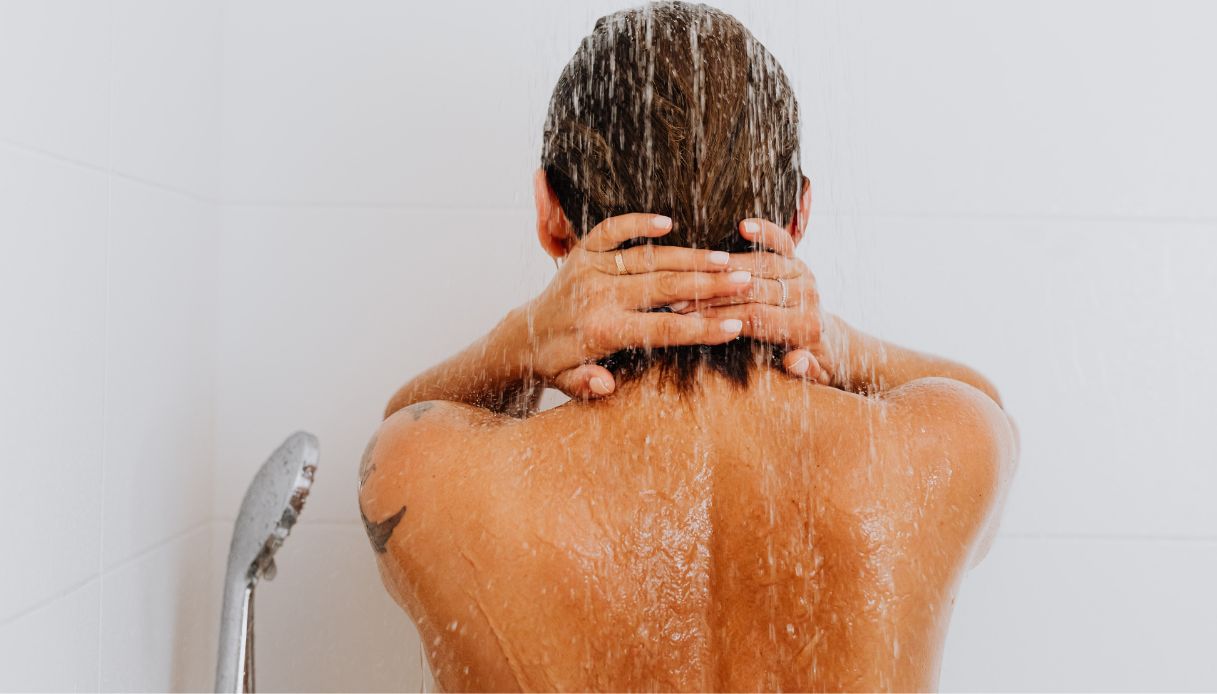 primo piano schiena donna abbronzata sotto la doccia con mani dietro il collo e capelli bagnati