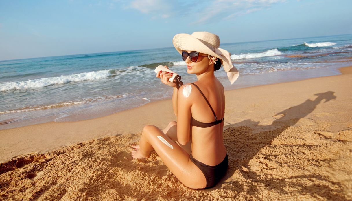 donna al mare con costume nero e cappello di paglia che applica crema solare sulla schiena con sfondo spiaggia e mare