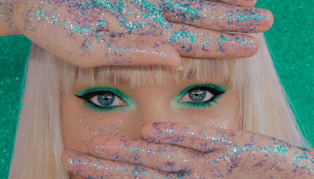 closeup occhi azzurri con lenti a contatto e trucco verde e faccia coperta da mani con glitter