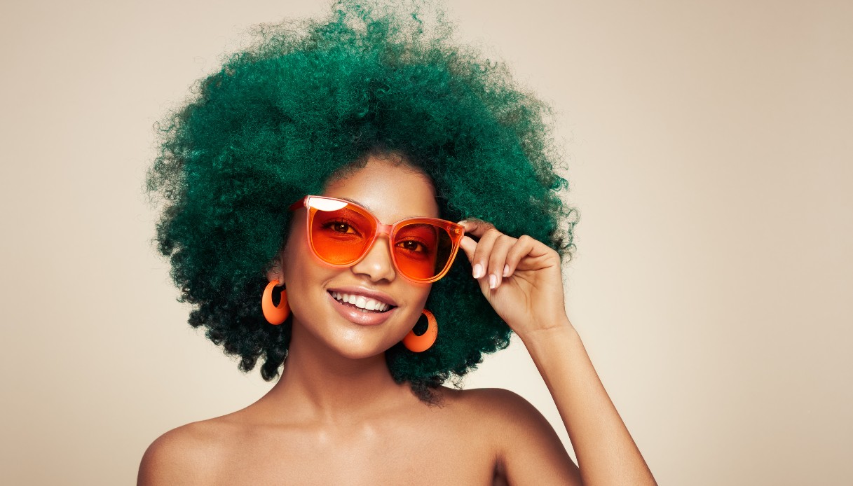 Una ragazza nera con i capelli ricci tinti di verse scuro indossa orecchini arancioni e occhiali da sole trasparenti con le lenti rosse