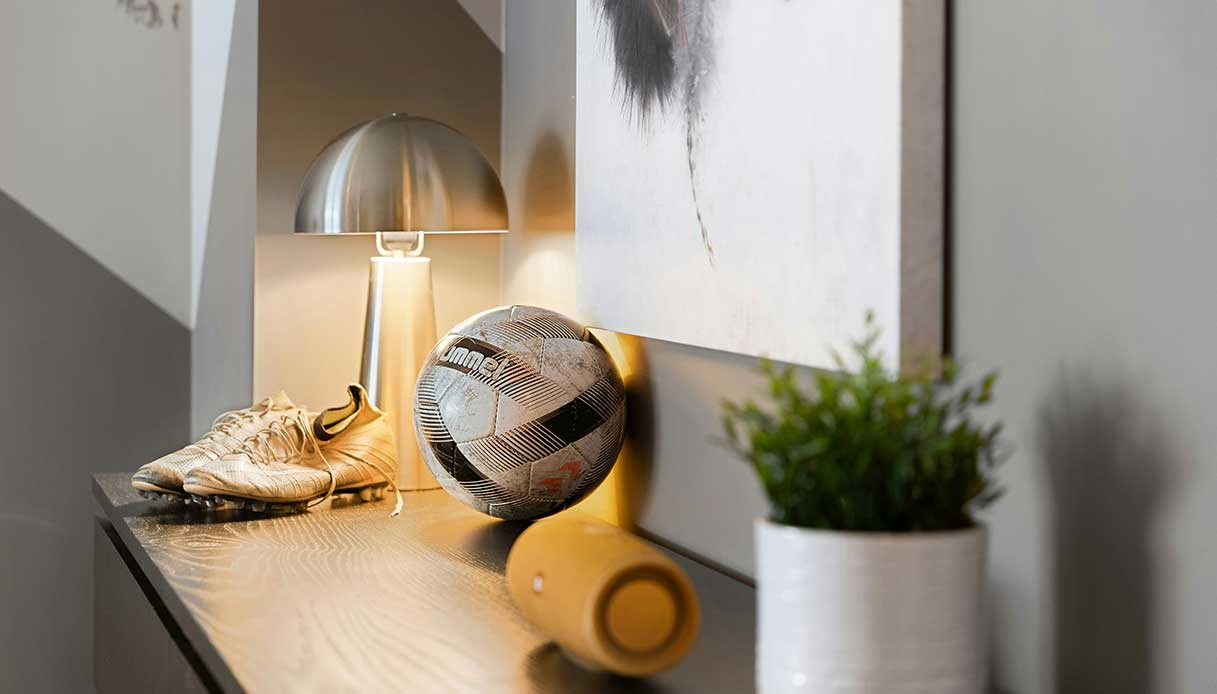 Ingresso con lampada da tavolo, scarpe da calcio, pallone e pianta in vaso su una mensola.