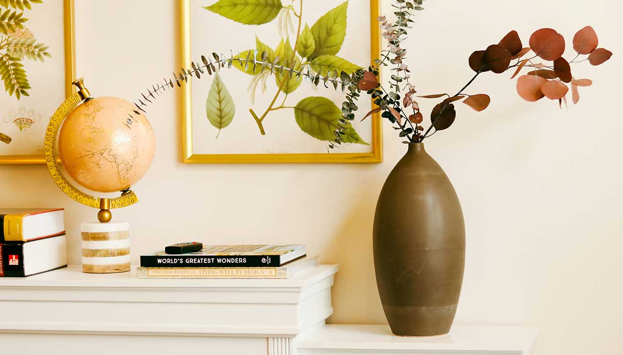 Dettaglio d’ingresso con vaso marrone contenente rami secchi, mappamondo e libri decorativi.