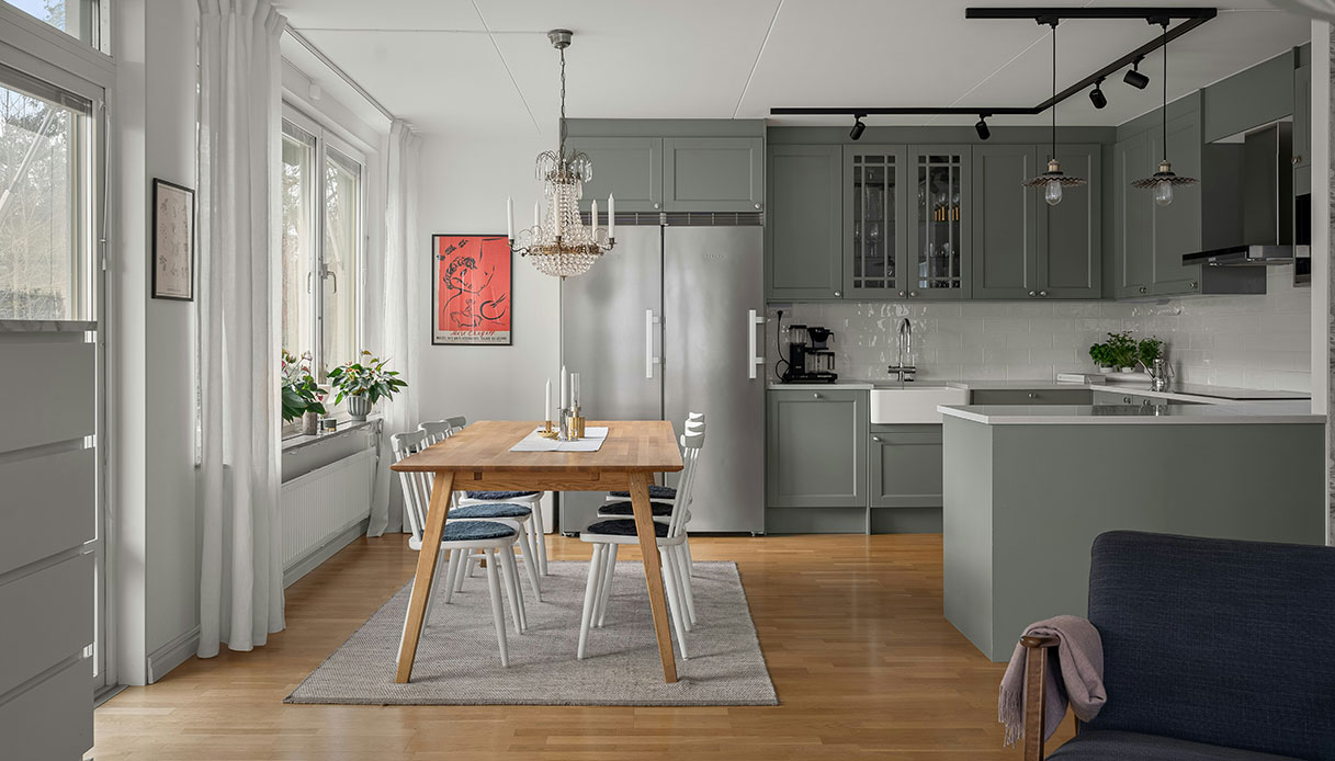 Cucina luminosa in stile nordico con mobili verde salvia, tavolo in legno e grandi finestre.