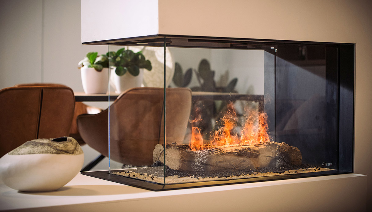Camino elettrico con effetto fiamma realistico inserito in una struttura in vetro trasparente, in un soggiorno moderno con arredi naturali.