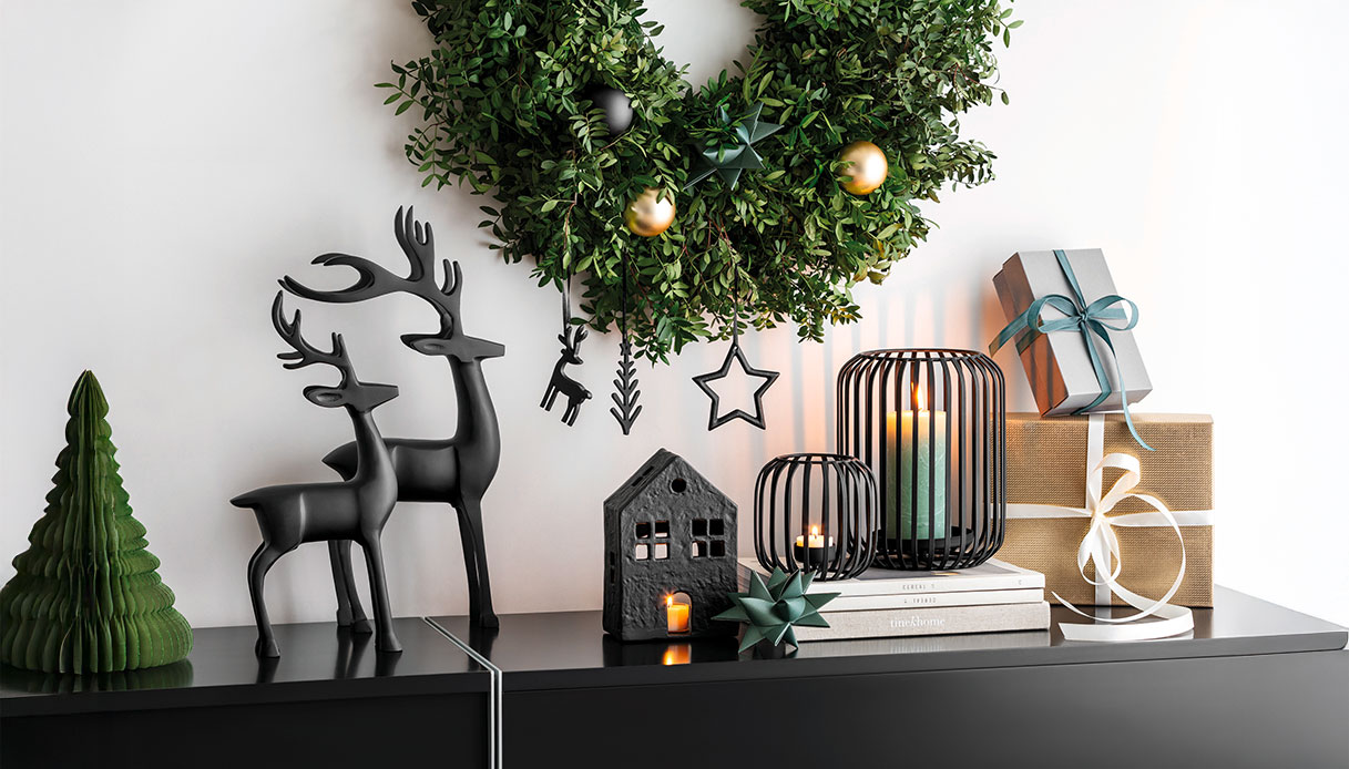 Decorazioni natalizie in nero opaco con renne stilizzate, lanterne e ghirlanda verde su mobile moderno.
