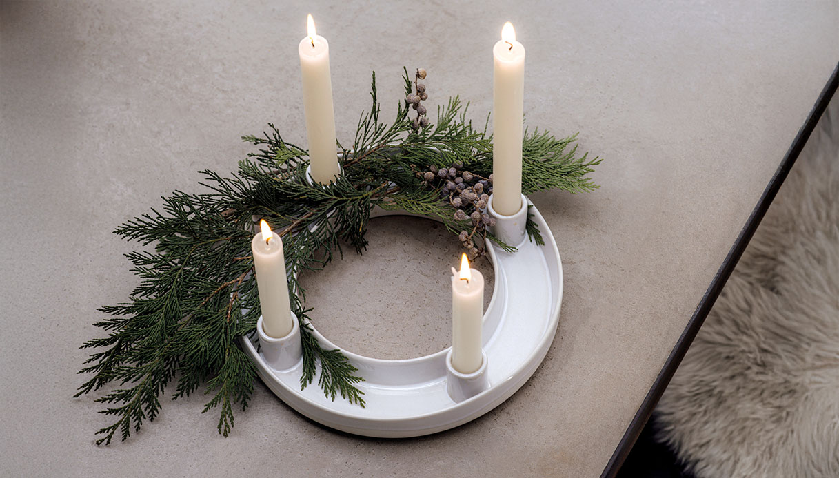 Corona dell&rsquo;Avvento minimalista con candele bianche e rami verdi su tavolo chiaro.