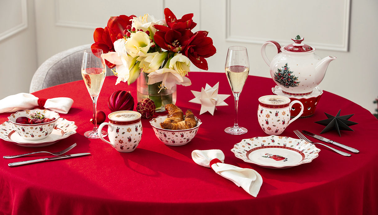 Colazione natalizia con servizi Toy’s Delight su tovaglia rossa e bouquet di fiori.