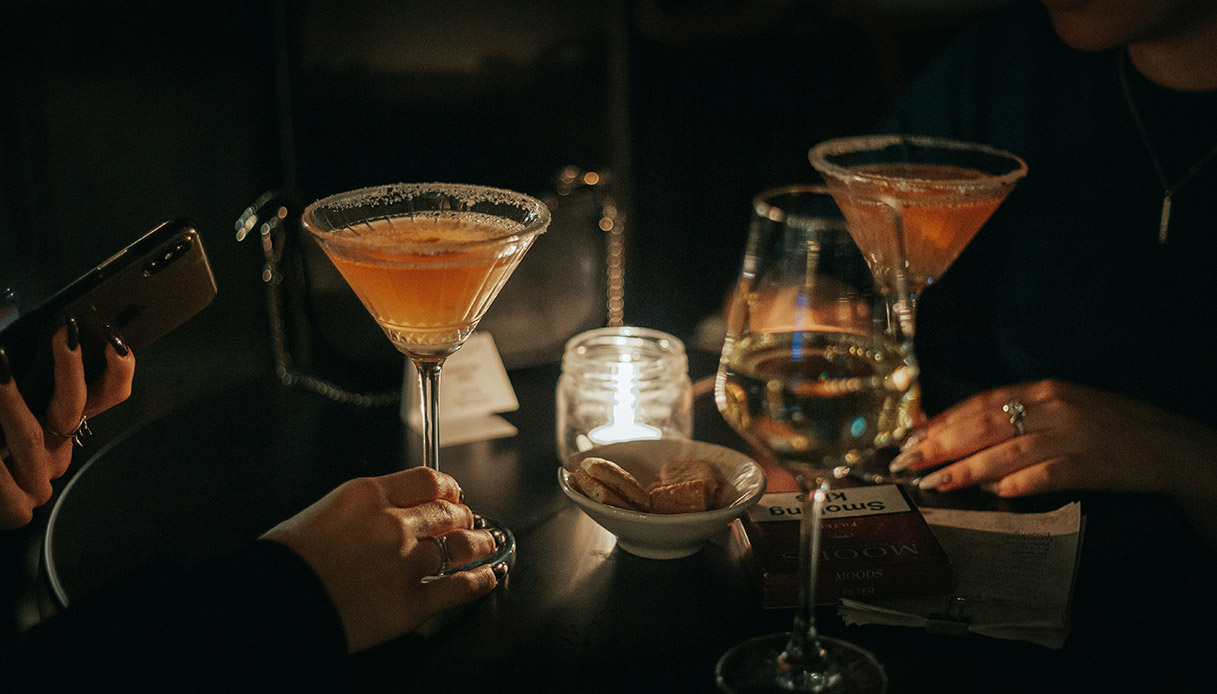Mani che sorreggono calici e cocktail in un ambiente soft, atmosfera conviviale da aperitivo serale