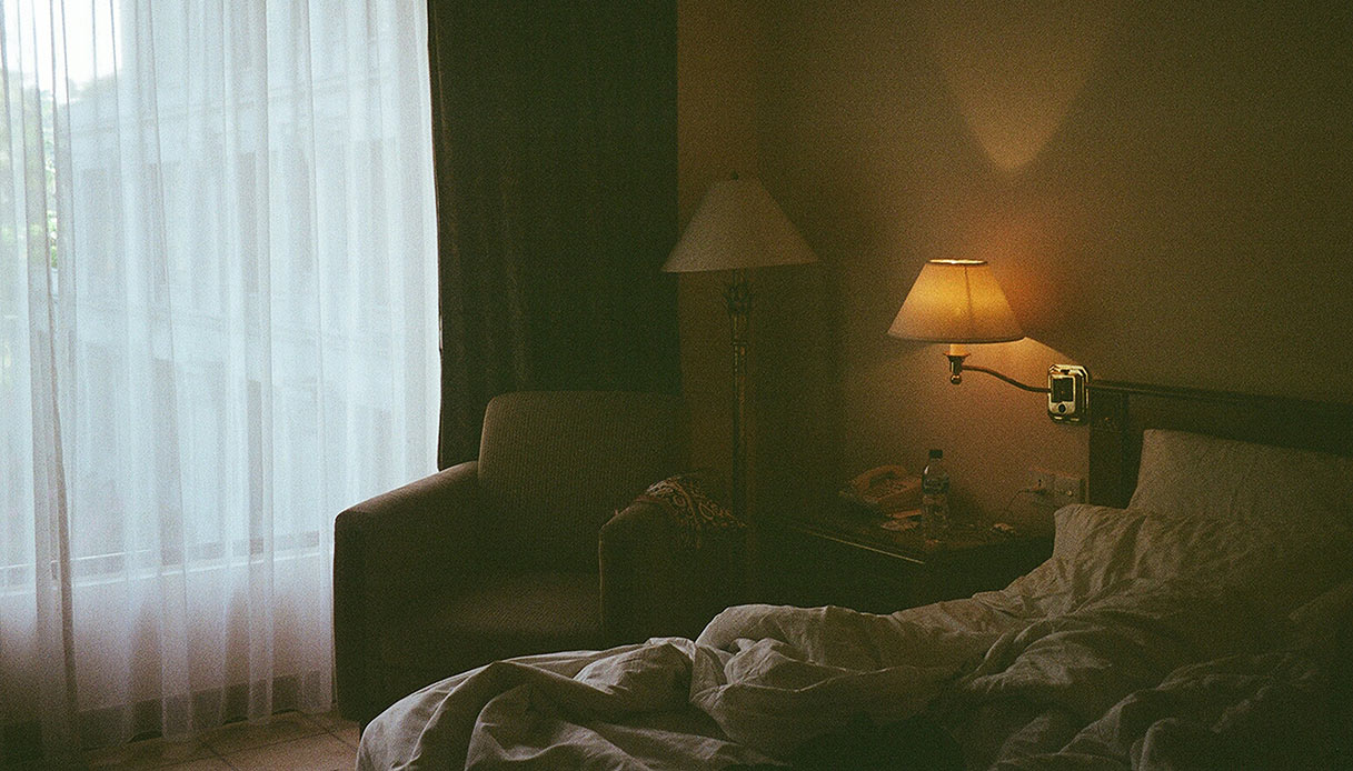 Camera con letto sfatto, poltrona accanto alla finestra e lampade che creano un&rsquo;atmosfera calda.