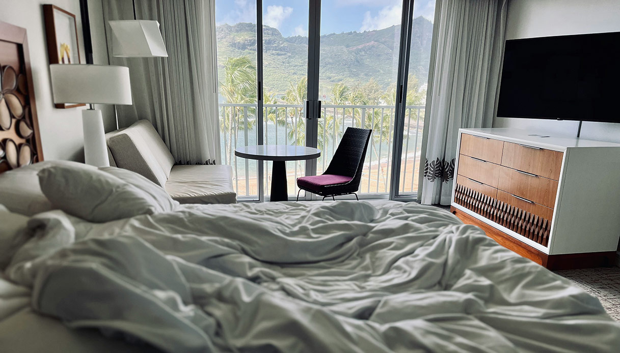 Camera con letto sfatto, tavolino e ampia finestra che si apre su palme e montagne.