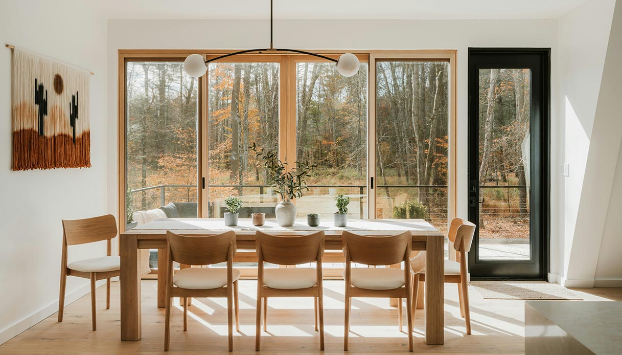 Zona pranzo in legno con grandi vetrate affacciate sul bosco in ambiente montano.