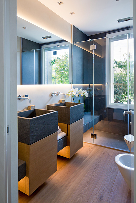 Bagno moderno con doppi lavabi in pietra scura, specchio retroilluminato, doccia in vetro e parquet in rovere