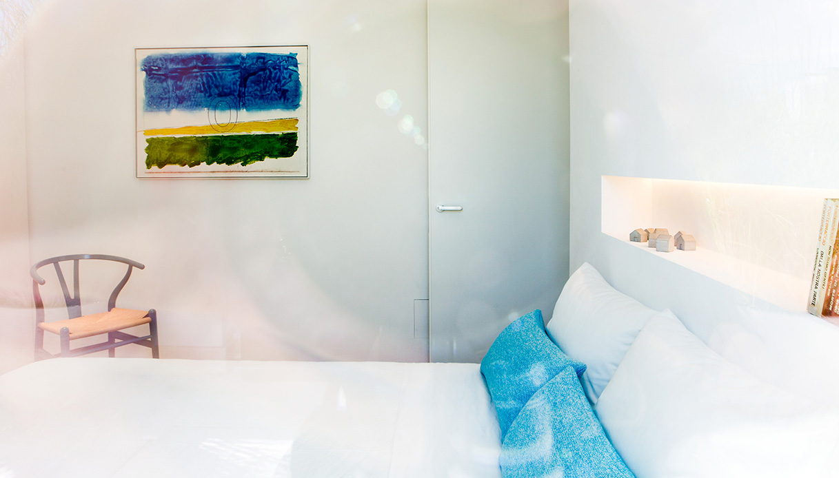 Camera da letto minimal con biancheria chiara, nicchia retroilluminata e quadro astratto blu e verde