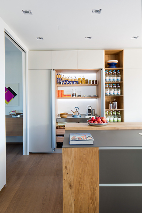 Cucina con dispensa nascosta retroilluminata, scaffali ordinati e bottiglie a vista accanto all’isola centrale