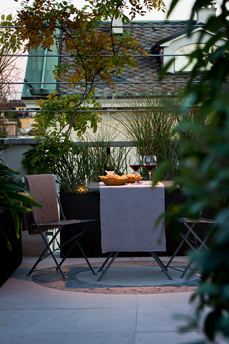 Terrazza dell’attico a Milano con tavolino apparecchiato, calici di vino e piante, vista sui tetti della città