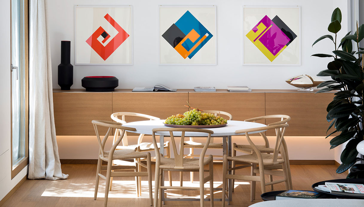 Zona pranzo dell’attico con tavolo rotondo bianco, sedie Wishbone in legno naturale e madia su misura con illuminazione LED