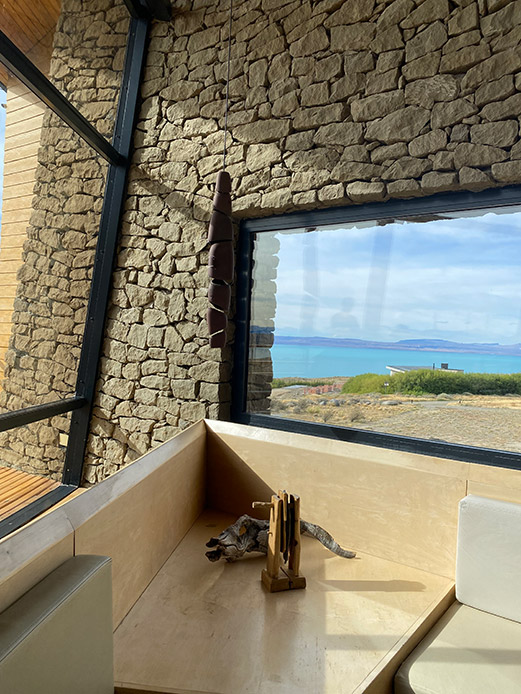 Interno con muro in pietra a vista e grande vetrata affacciata su lago e paesaggio arido patagonico