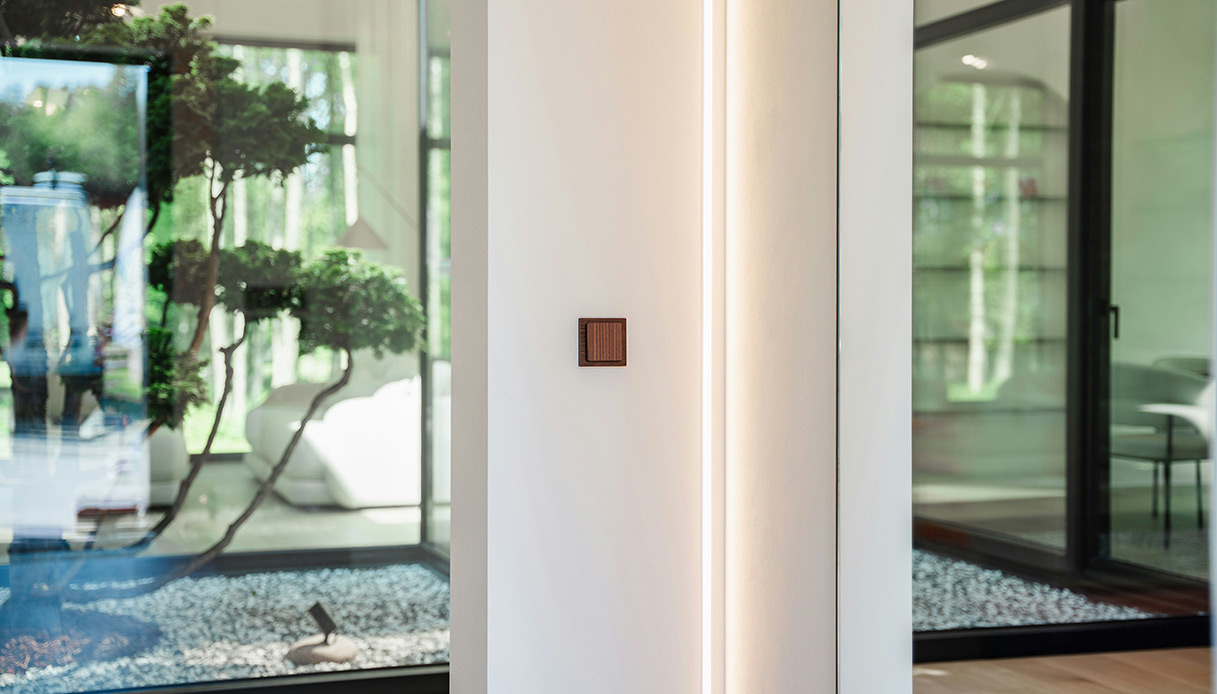 Interruttore domotico in legno su parete bianca con striscia LED verticale, in un interno contemporaneo con vetrate su giardino