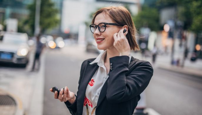 Una donna con gli occhiali sorride in una strada di citt&agrave; mentre tocca un auricolare nero nell'orecchio e tiene la custodia nell'altra mano.