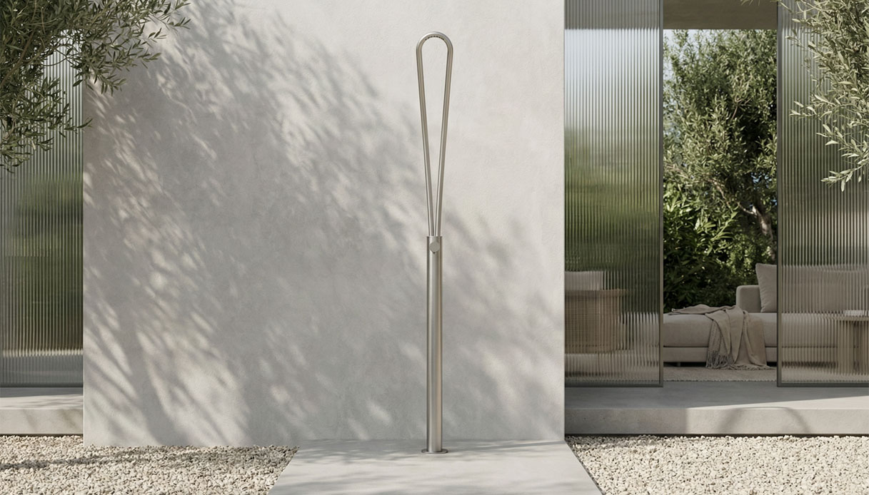 Colonna doccia outdoor Cruna in acciaio inox satinato su pavimento in pietra chiara, con ulivi sullo sfondo