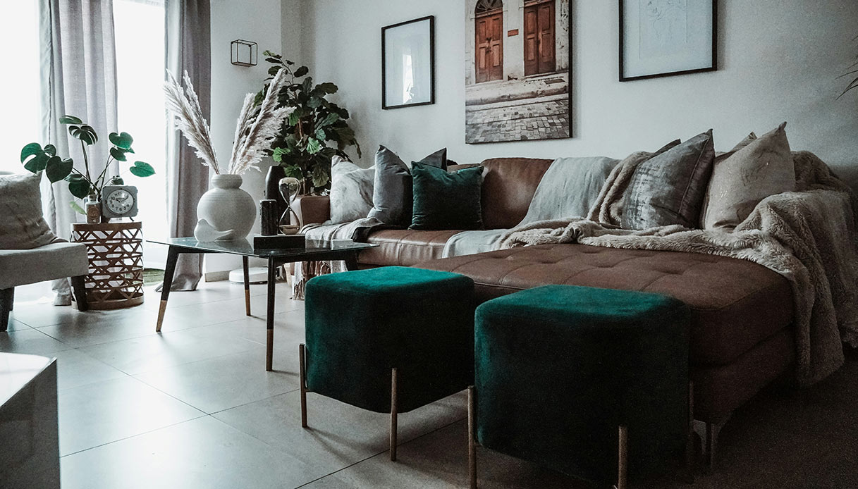 Soggiorno con divano in pelle marrone, due poggiapiedi quadrati in velluto verde smeraldo con gambe in legno, tavolino in vetro e piante d'appartamento
