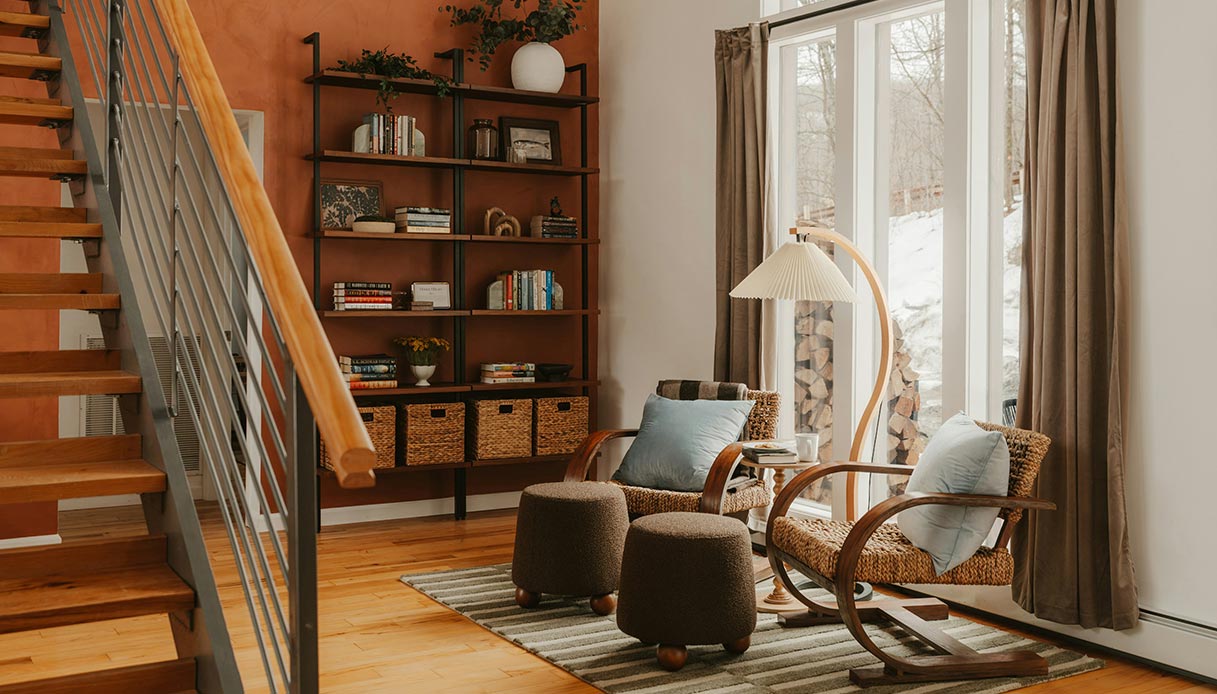 Angolo soggiorno con due pouf rotondi in boucl&eacute; marrone, poltrone in rattan con cuscini azzurri, lampada ad arco in legno e libreria a parete color terracotta