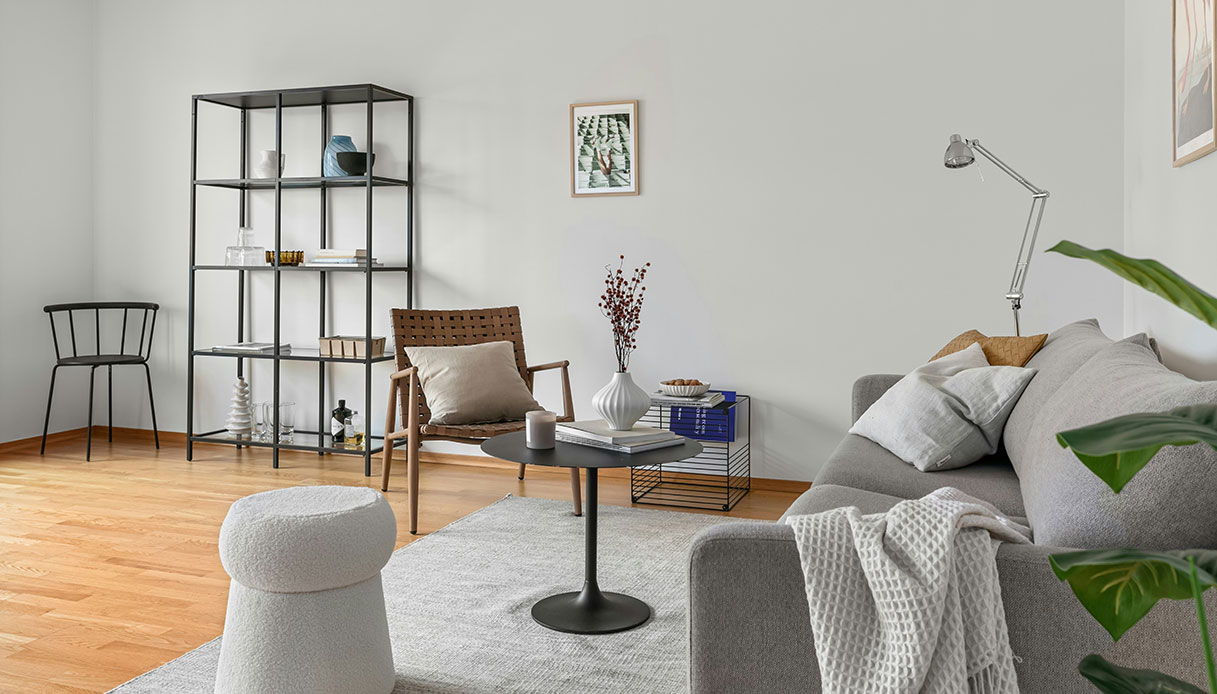 Soggiorno scandinavo con pareti bianche, divano grigio, pouf tondo in boucl&eacute; bianco, poltrona in pelle intrecciata, scaffale in metallo nero e parquet in legno chiaro