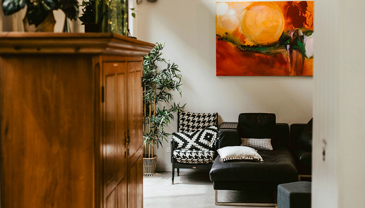 Soggiorno eclettico con chaise longue in pelle nera, poltrona in bianco e nero, mobile antico in legno, pianta di bamb&ugrave; e dipinto astratto arancione sulla parete