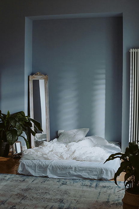 Camera da letto con pareti azzurro polvere, letto basso con biancheria bianca, specchio vintage con cornice dorata a pavimento e piante tropicali verdi ai lati