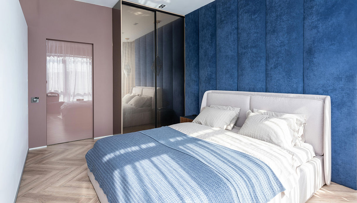 Camera da letto moderna con parete imbottita in velluto blu intenso, letto con testiera bianca, biancheria bianca e blu, parquet a spina di pesce e porta color rosa antico