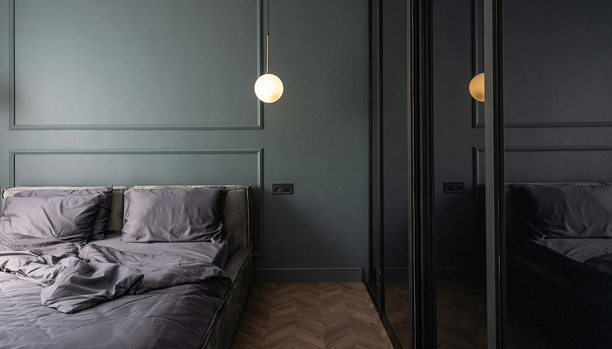 Camera da letto con pareti verde acqua scuro e modanature, letto basso con biancheria grigia, lampade a sospensione con sfera in vetro bianco e specchio a parete