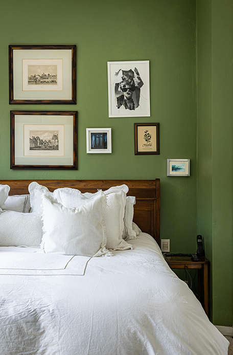 Camera da letto con pareti verde salvia, testiera in legno massello scuro, biancheria bianca e composizione di stampe incorniciate sulla parete