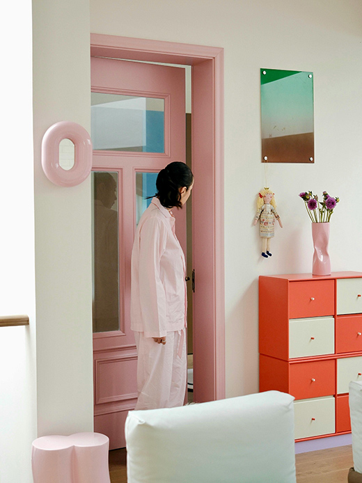 Interno con porta verniciata rosa, cassettiera modulare arancione e bianca, vaso rosa con fiori viola e scultura da parete rosa