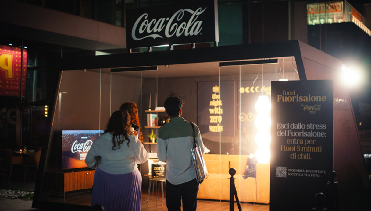 Due visitatori di spalle davanti al pop-up Coca-Cola Glass House di notte, struttura con pareti trasparenti illuminate e insegna Coca-Cola in alto, cartello "Fuori Fuorisalone" con QR code