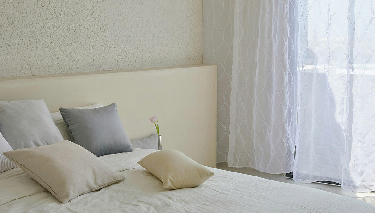 Camera da letto cocoon style con tende bianche che filtrano la luce e cuscini neutri