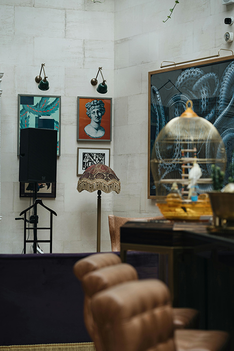 Interno eclettico con stampe di busti classici a parete, lampada a stelo con paralume d'epoca, gabbia dorata e poltrona in pelle marrone in primo piano