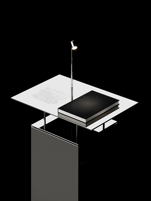 Leggio cromato con libro aperto e piccola lampada da lettura su piedistallo metallico, installazione Jil Sander Reference Library Milano Design Week 2026